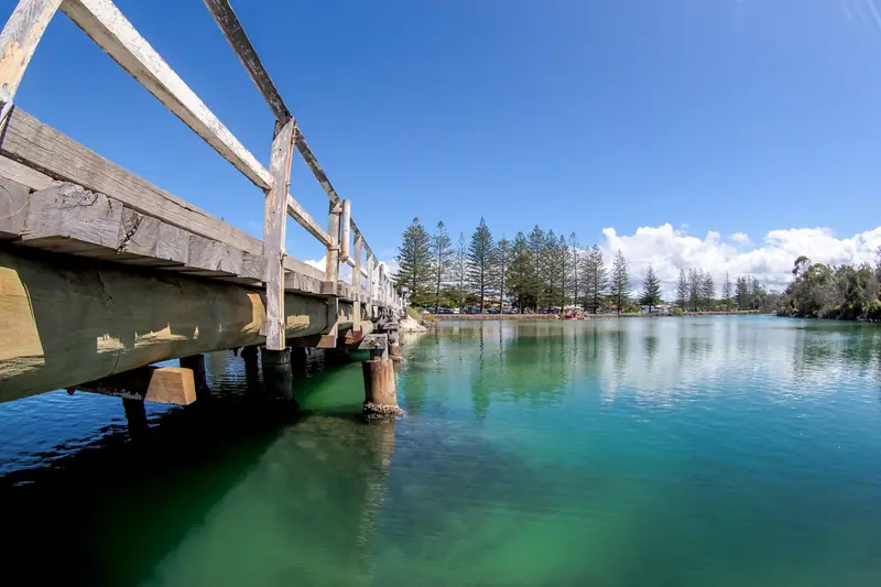 Timber footbridge over clear green water with Norfolk pines on the foreshore
